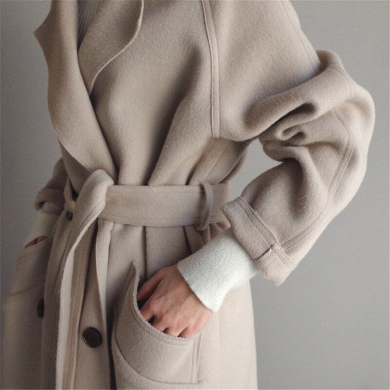 Beige uldfrakke med bælte og store lommer, elegant design, kvinde iført hvid sweater. Vintermode, stilfuldt overtøj, minimalistisk look.