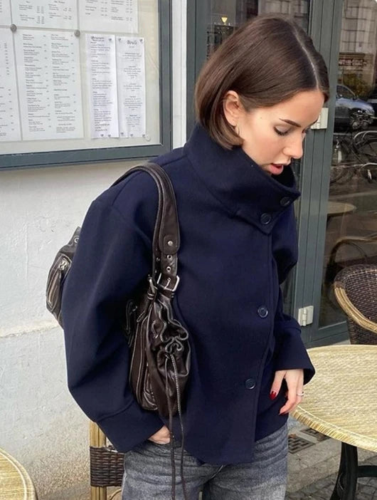 Kvinde i mørkeblå jakke med kort brunt hår, bærer en brun lædertaske. Står ved cafébord med menukort i baggrunden. Mode, efterår, byliv.