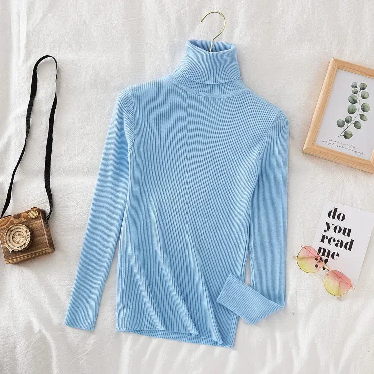 Lyseblå rullekrave sweater i ribstrik på hvid baggrund, ved siden af et vintage kamera, solbriller og en ramme med planteillustration.