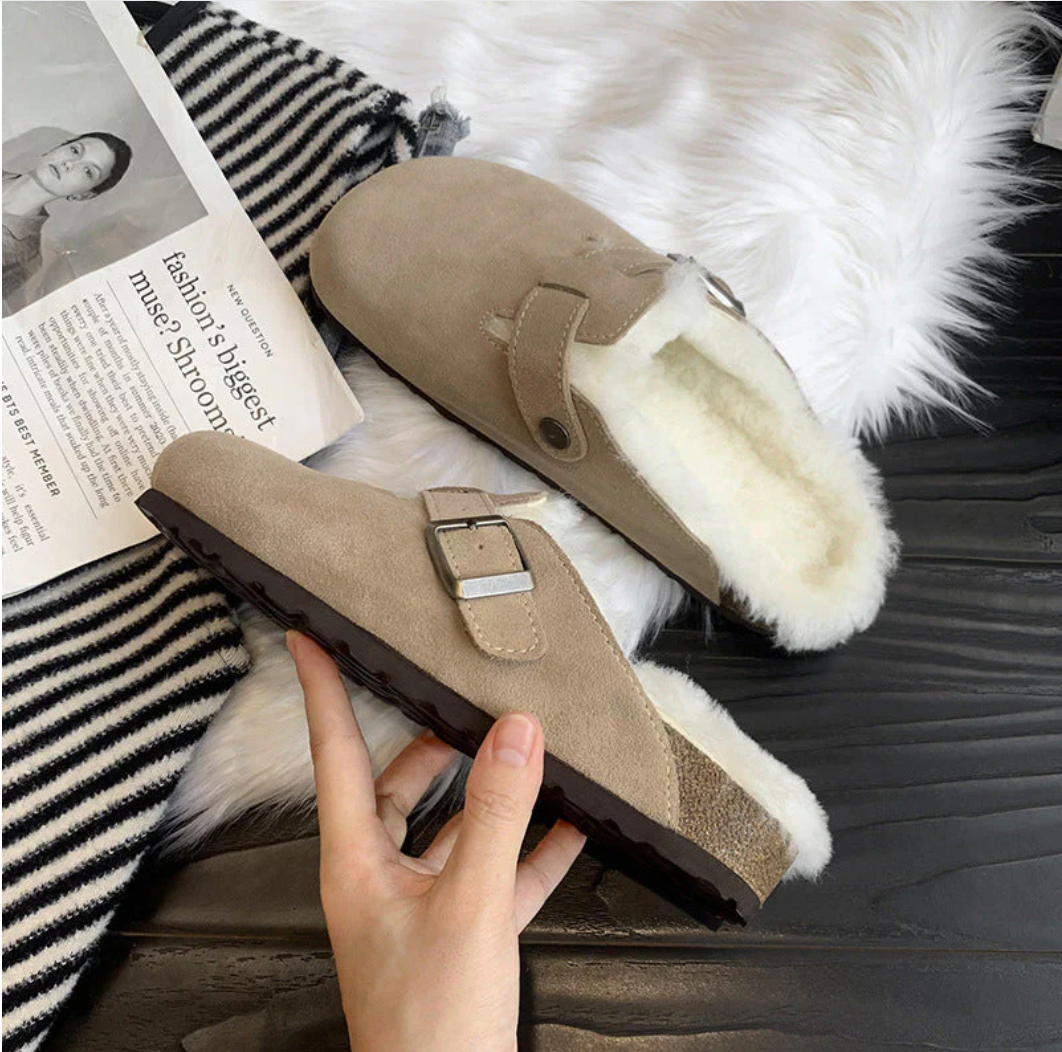 Beige hjemmesko med pelsfor, spænde og skridsikker sål på sort træbord, ved siden af stribet tæppe og modeblad. Komfortable vinterfodtøj.