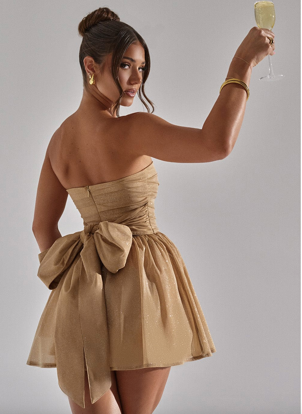 Kvinde i gylden cocktailkjole med stor sløjfe, holder et glas champagne. Elegant festlook, perfekt til gallafest eller nytårsaften.