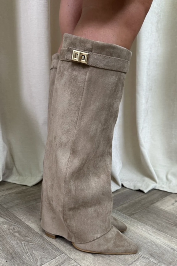 Beige ruskindsstøvle med guldspænde, stående på trægulv foran hvidt gardin. Elegant fodtøj, moderne design, efterårsmode.