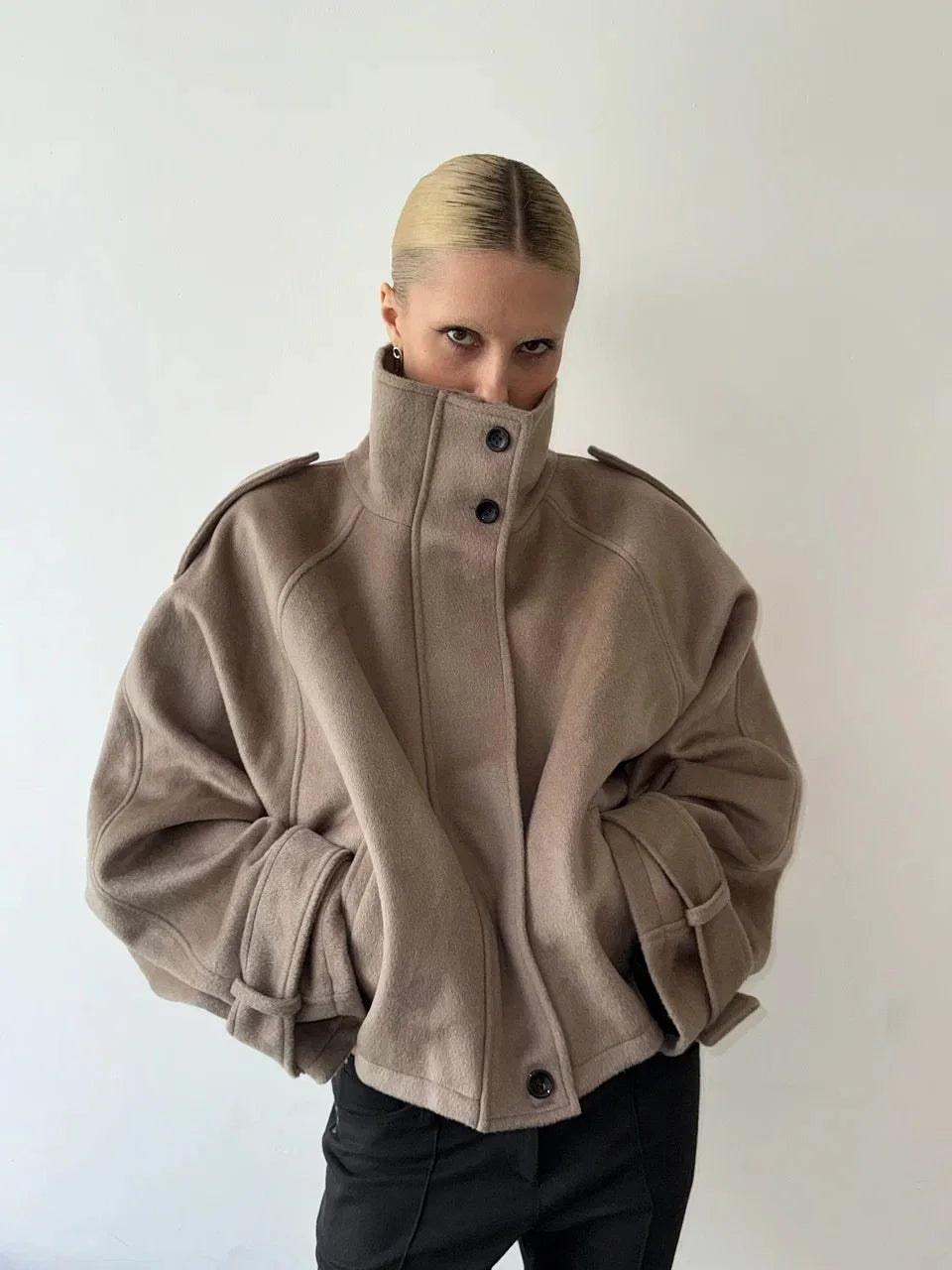 Kvinde iført beige oversized frakke med høj krave og sorte knapper, poserer mod hvid baggrund. Trendy vintermode, minimalistisk stil.