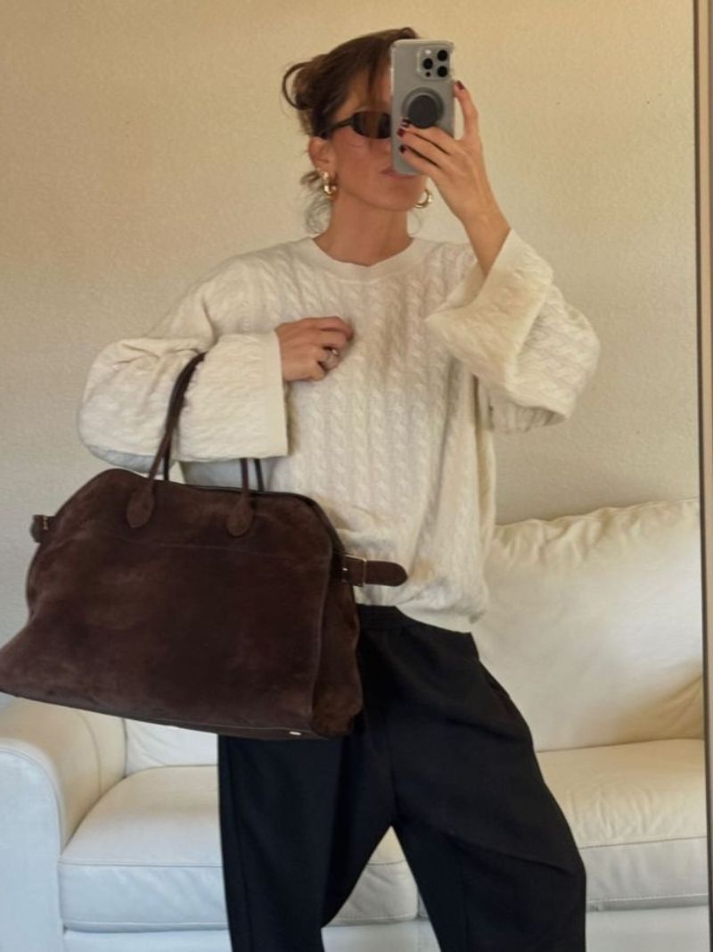 Kvinde i hvid sweater og sorte bukser tager selfie med stor brun taske. Hun bærer solbriller og guldøreringe, står foran hvid sofa.