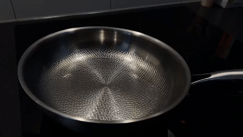 Rustfrit stål stegepande med bikagemønster på sort komfur. Perfekt til jævn varmefordeling og non-stick madlavning. Ideel til køkkenentusiaster.