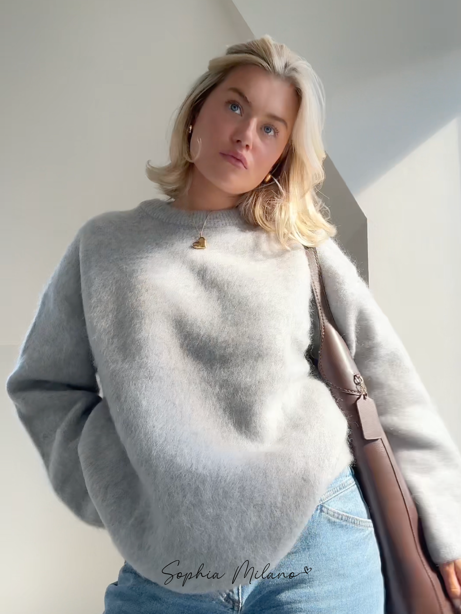 Ung kvinde i grå sweater og jeans, bærer brun taske. Moderne mode, afslappet stil, naturligt lys. Perfekt til efterårsgarderobe.