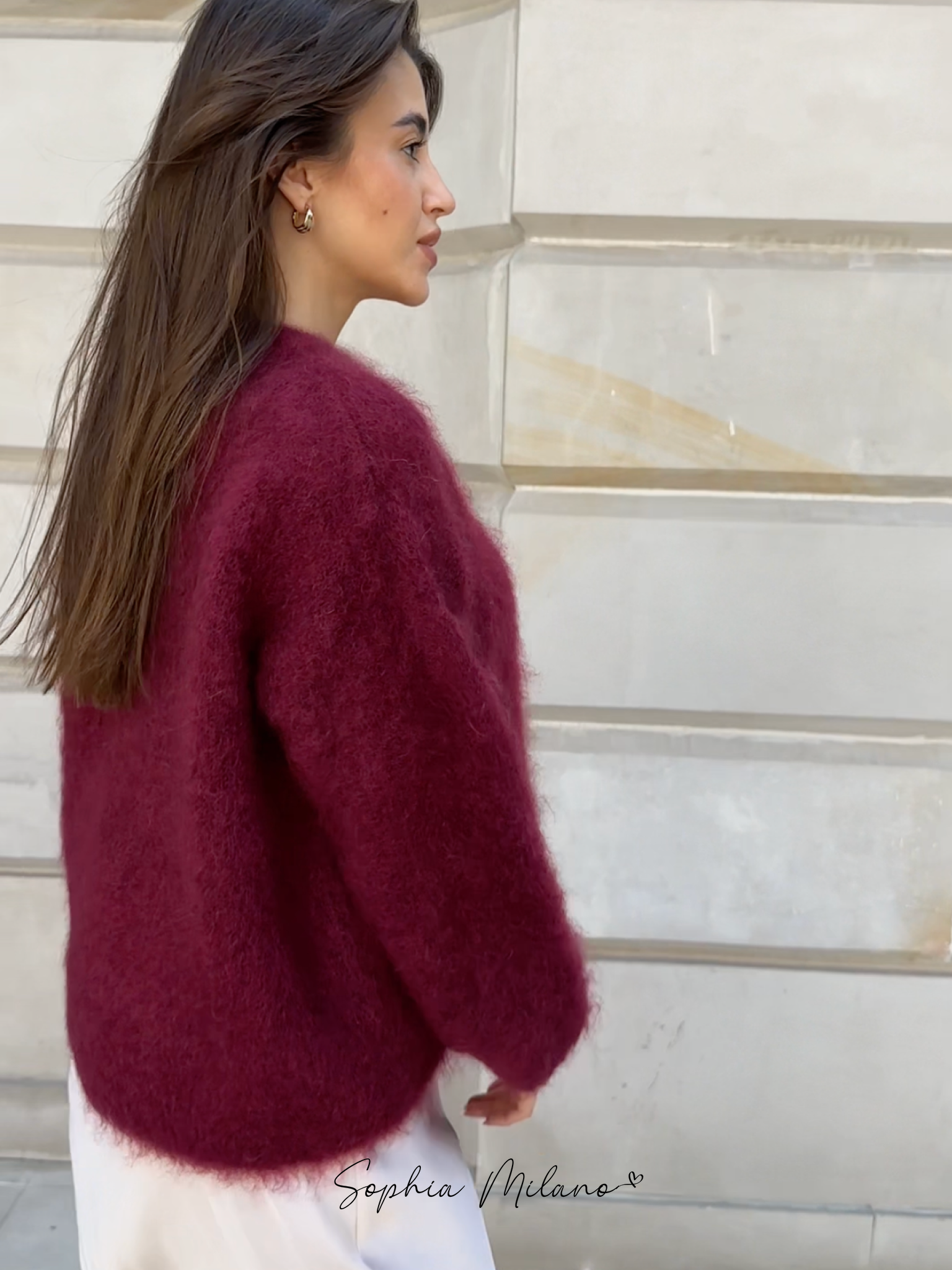 Kvinde iført bordeauxrød mohairtrøje, set fra siden, mod beige murstensvæg. Langt brunt hår, elegant og stilfuldt look.