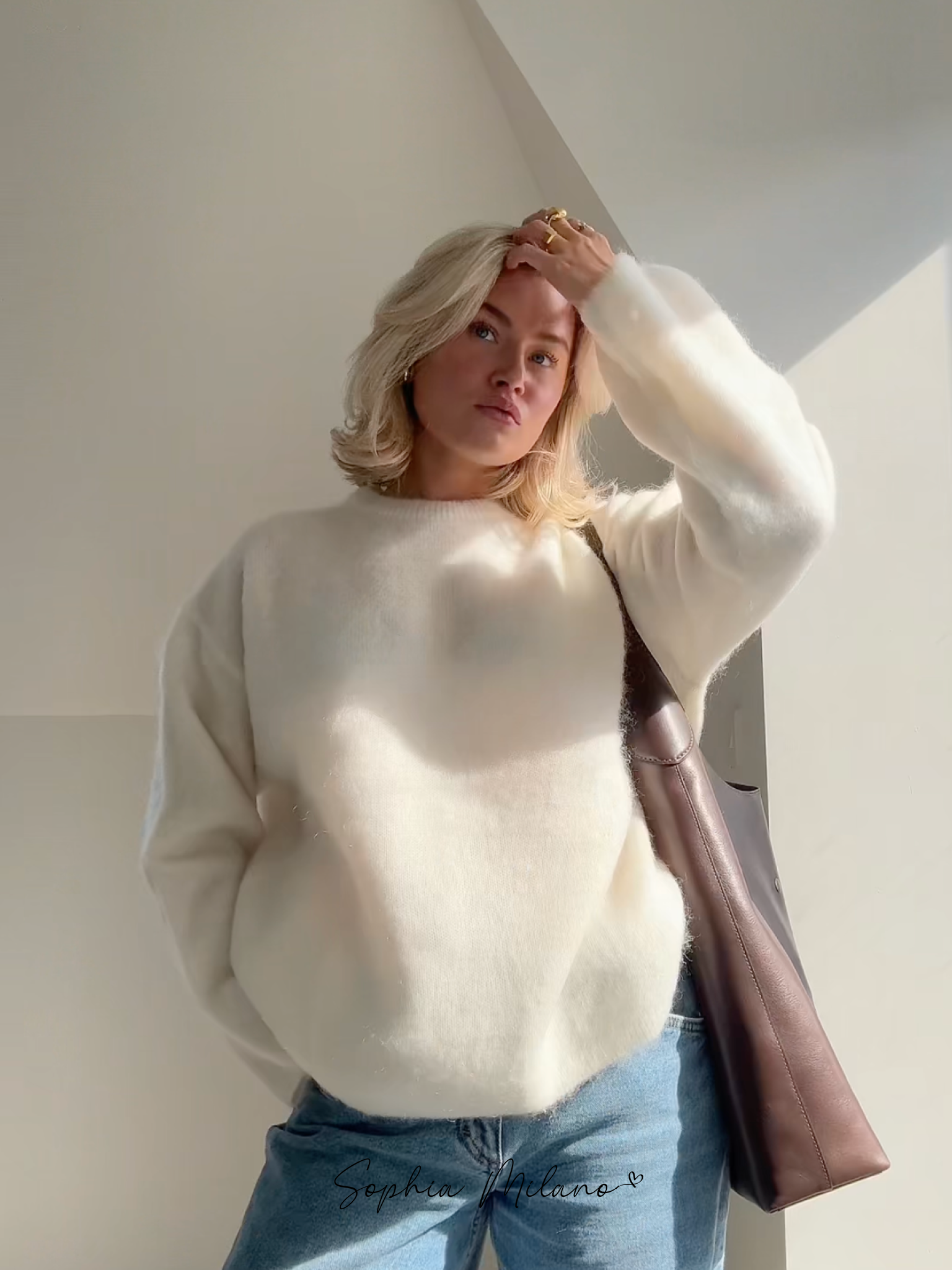 Ung kvinde i hvid sweater og jeans, poserer i naturligt lys. Hun har en brun taske over skulderen. Moderne mode, afslappet stil, skandinavisk design.