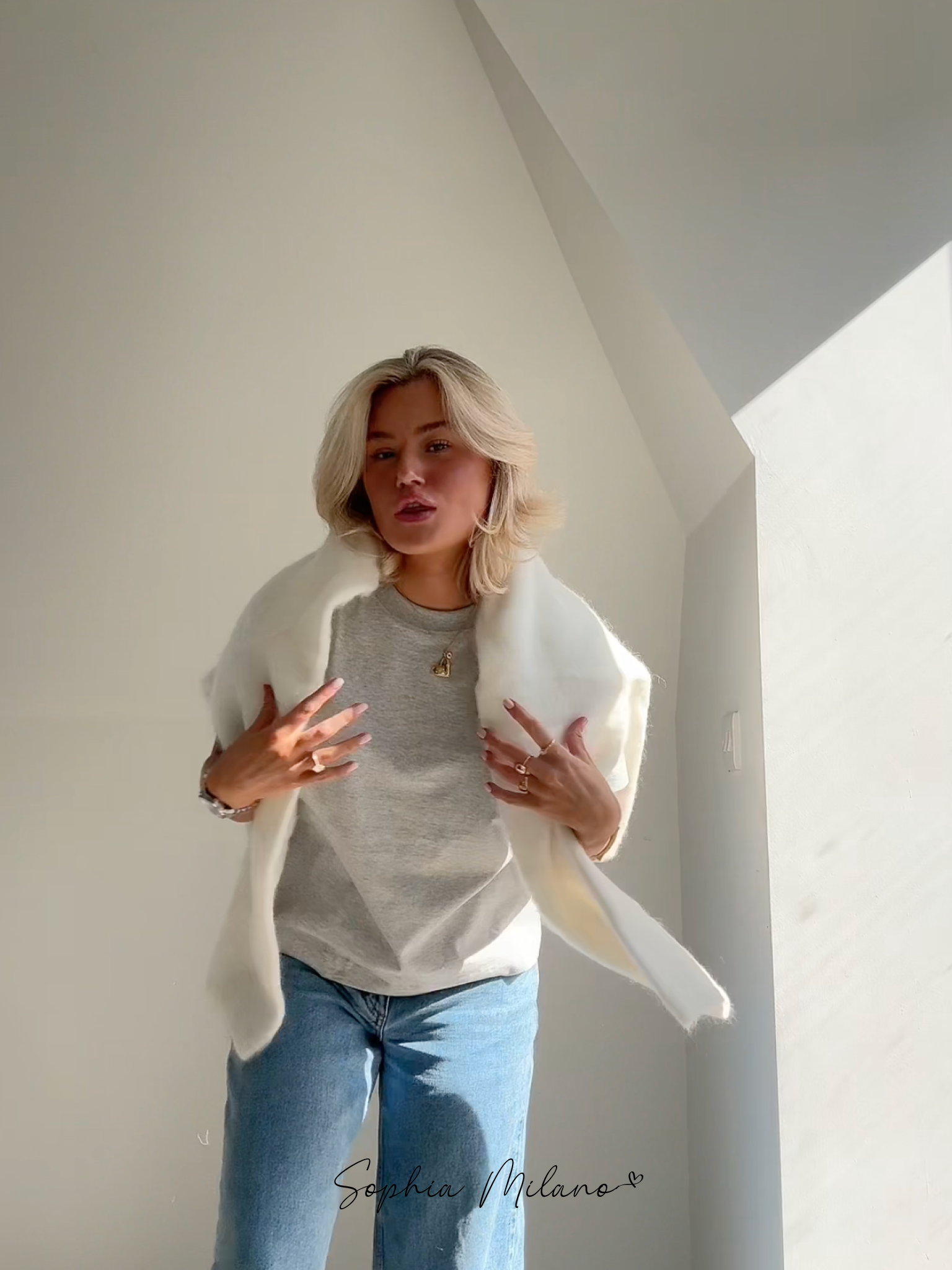 Ung kvinde i grå t-shirt og jeans, iført hvid pelsjakke, står i sollys ved hvid væg. Mode, stil, moderne kvindetøj, minimalistisk look.