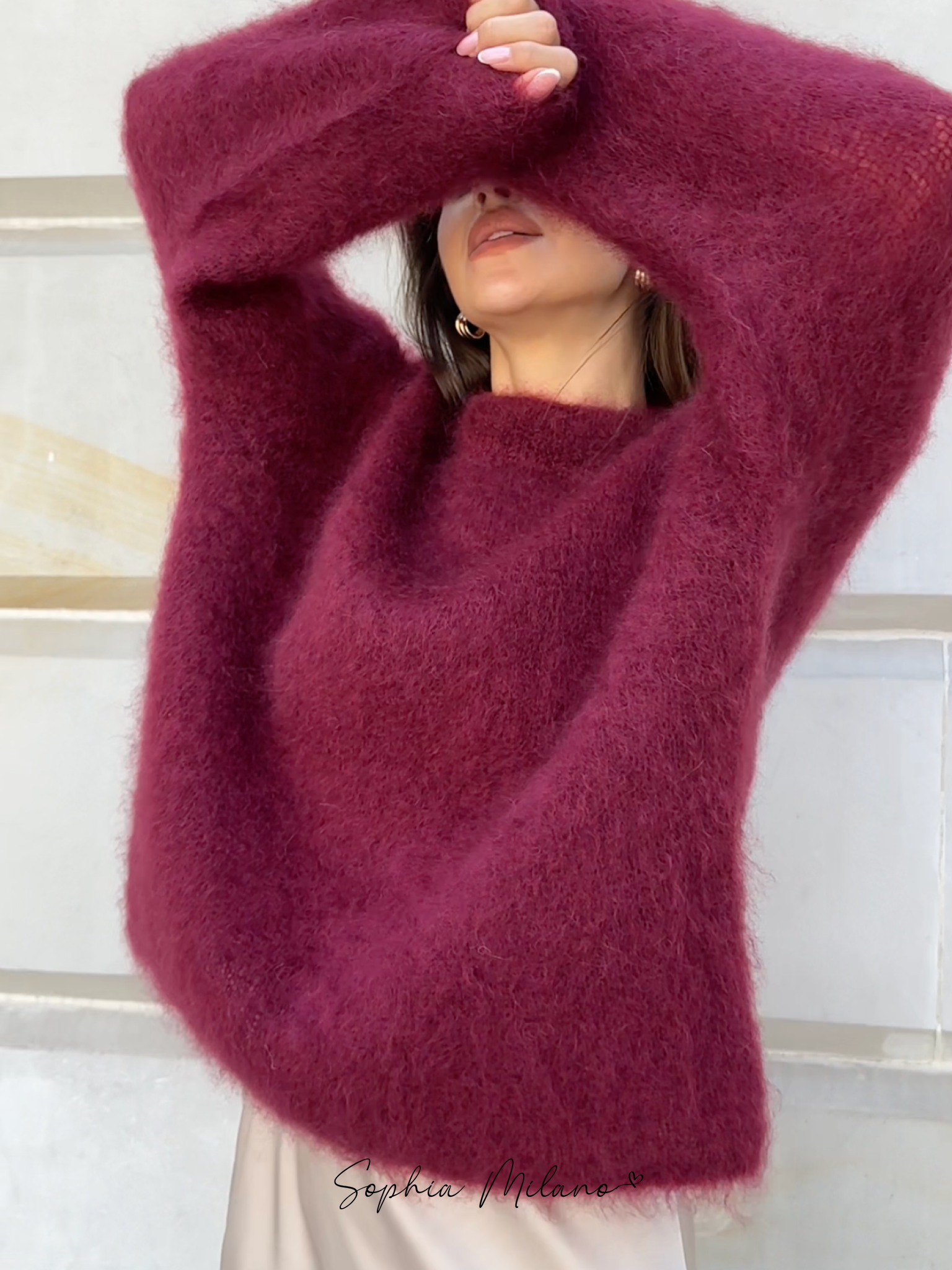 Kvinde iført en blød, bordeaux striktrøje fra Sophia Milano, poserer foran en lys væg. Perfekt til efterårsmode og stilfulde vinteroutfits.