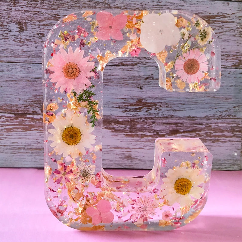 Bogstav C i harpiks med indlejrede tørrede blomster, herunder margueritter og lyserøde blomster, på en rustik træbaggrund. Dekorativt og unikt design.