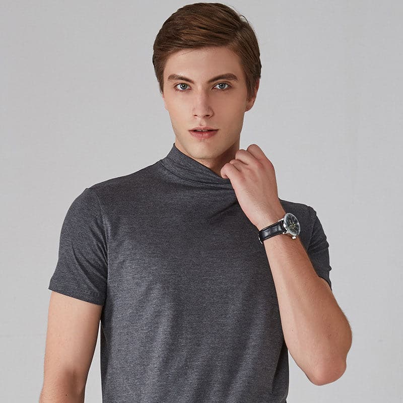 Jeg ved ikke, hvem personen er, men her er en beskrivelse: Mand i grå t-shirt med ur, ser direkte på kameraet. Moderne herremode, stilfuldt look.