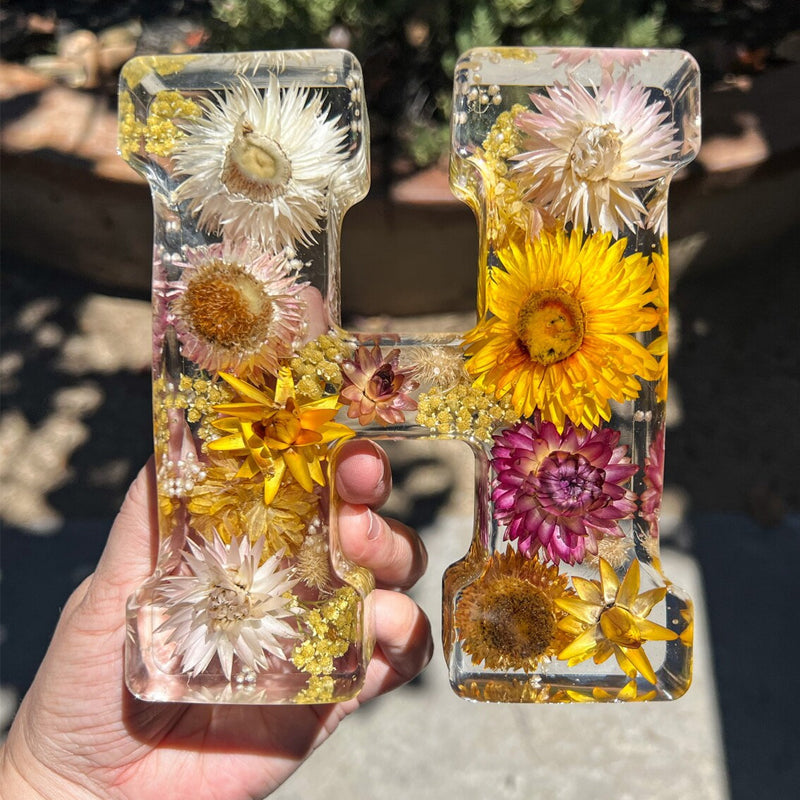 Hånd holder et gennemsigtigt harpiksbogstav "H" fyldt med tørrede blomster i gule, hvide og lilla nuancer. Perfekt til boligindretning og kunsthåndværk.