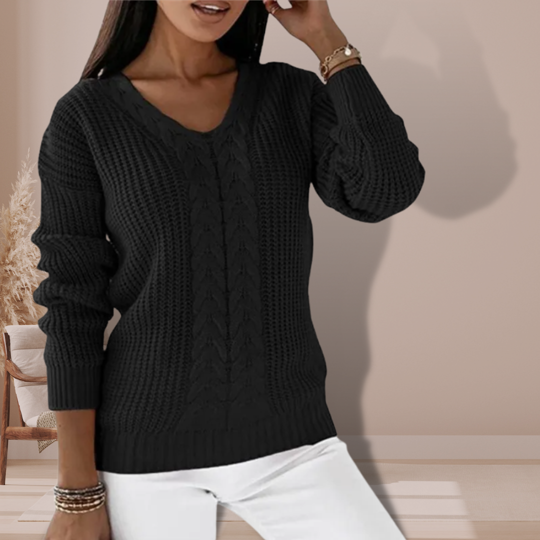 Kvinde iført sort striktrøje med V-hals og hvid bukser, står i moderne indretning. Elegant efterårslook, mode til kvinder, stilfuldt outfit.