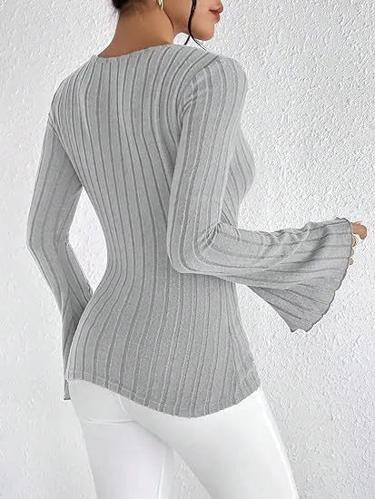 Kvinde iført grå ribstrikket trøje med klokkeærmer og hvide bukser, set fra siden mod hvid baggrund. Moderne mode, stilfuldt outfit.