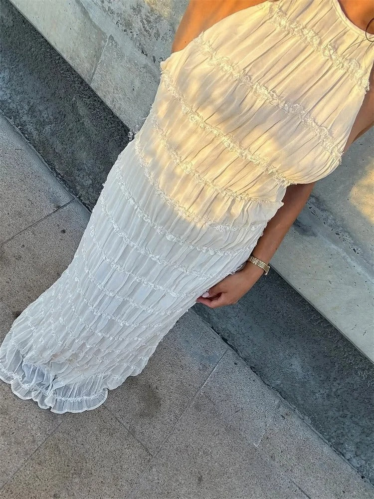 Kvinde i hvid, ærmeløs, plisseret kjole står op ad en grå væg. Elegant sommerkjole, perfekt til festlige lejligheder.