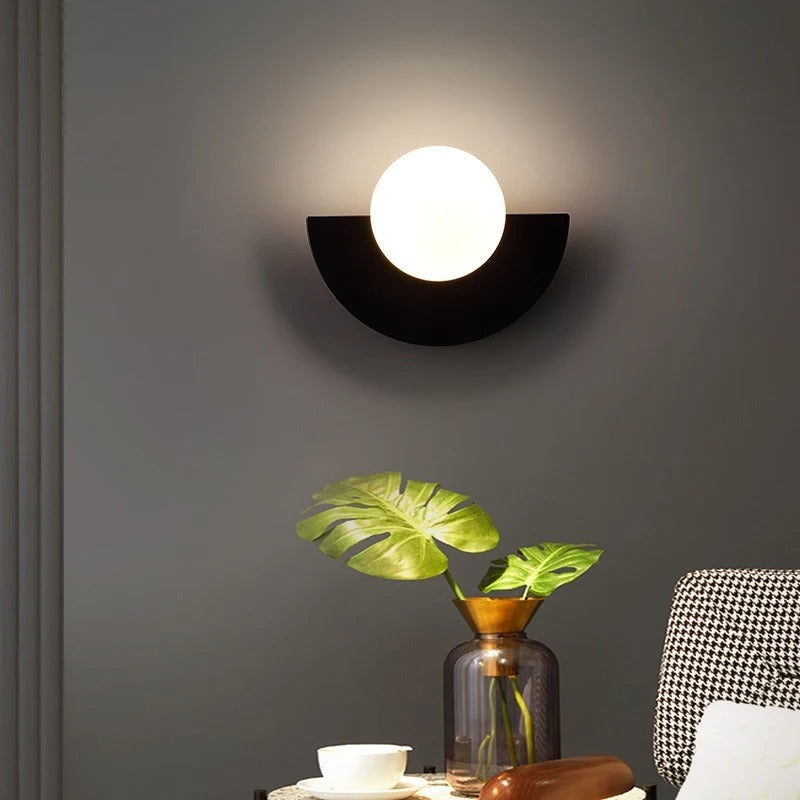 Moderne væglampe med halvmåneformet design og rund lyskilde, placeret over en glasvase med grønne blade, i stilfuldt interiør.