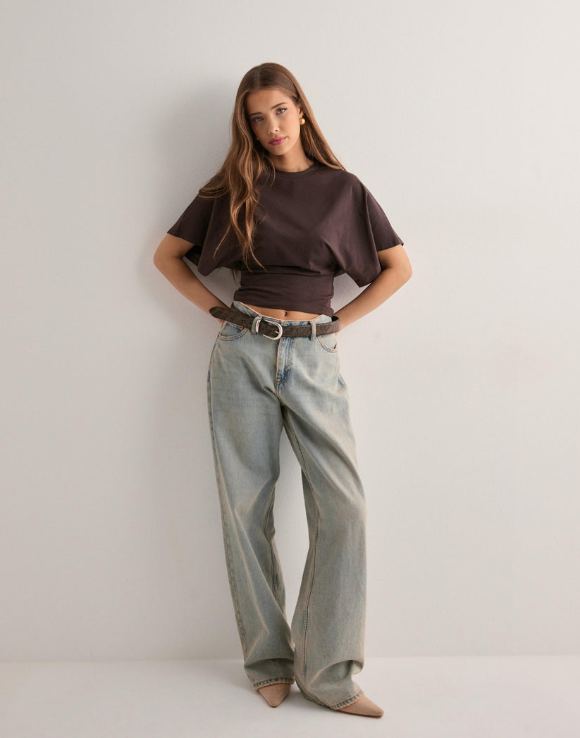 Ung kvinde i brun t-shirt og lyseblå jeans, poserer mod hvid baggrund. Mode, casual stil, moderne tøj, afslappet look, trendy outfit.