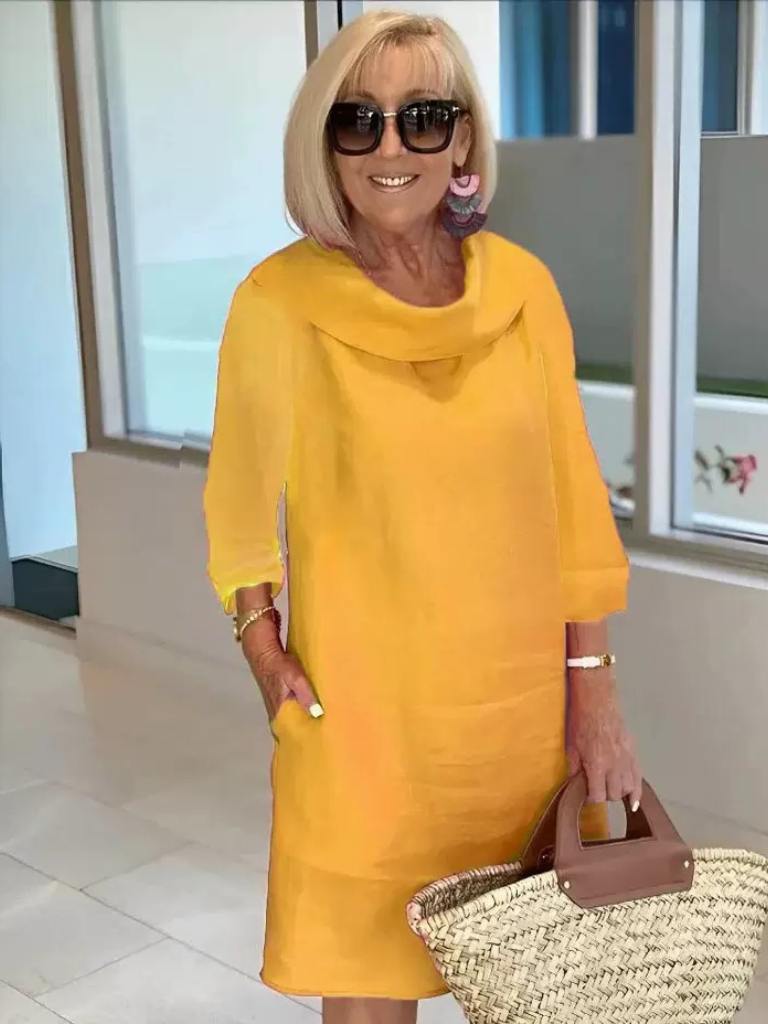 Kvinde i gul kjole med solbriller og øreringe, holder en stråtaske. Moderne mode, sommerstil, indendørs baggrund.