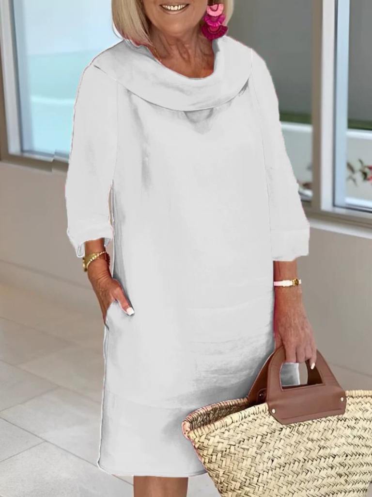 Ældre kvinde i hvid kjole med trekvartærmer, holder en stråtaske. Hun har lyserøde øreringe og står i et moderne, lyst rum.