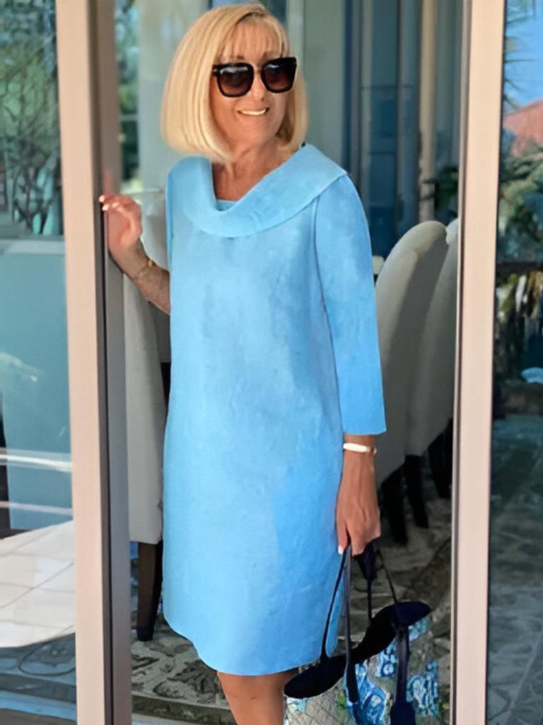 Kvindelig model i lyseblå kjole med solbriller, står ved glasdør. Sommermode, elegant stil, moderne kvindetøj, lyse farver.