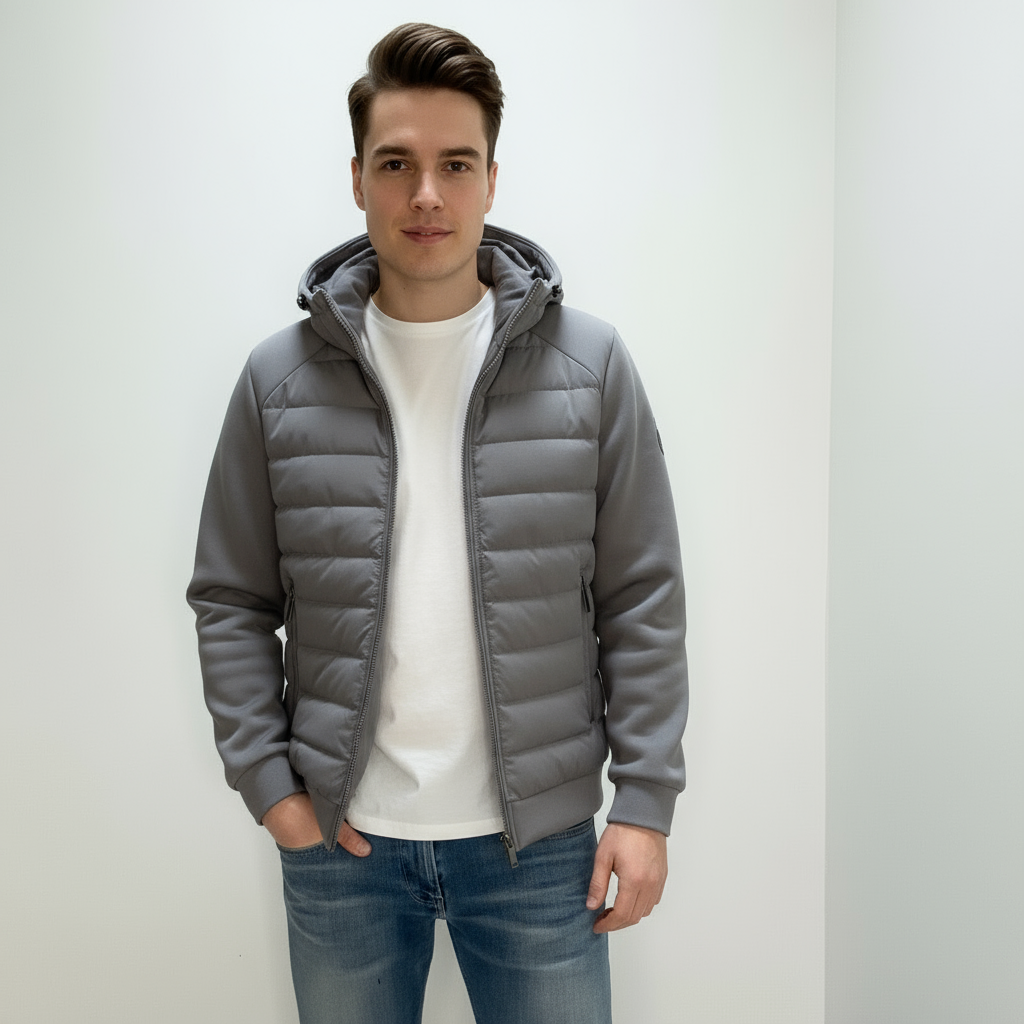 En mand iført en grå, quiltet jakke med hætte og en hvid t-shirt, står foran en lys baggrund. Casual mode, herretøj, moderne stil.