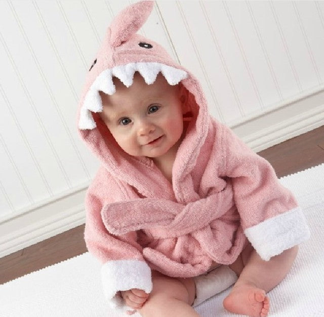 Baby iført lyserød hajbadekåbe med hætte og tænder, sidder på hvidt tæppe. Sødt babytøj, sjovt design, perfekt til badetid.