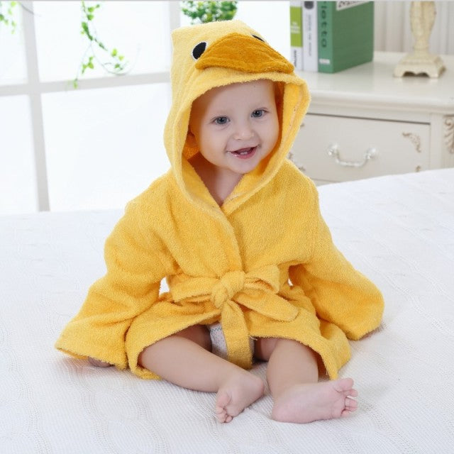 Baby iført gul ande-badekåbe sidder smilende på en seng i et lyst værelse. Sødt babybillede, børnetøj, badehåndklæde, indendørs.