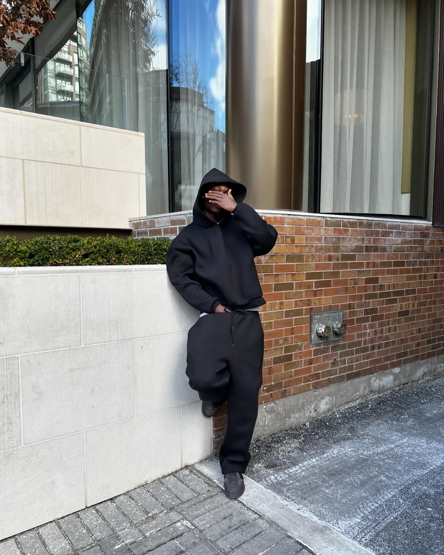 Mand i sort hættetrøje og joggingbukser læner sig op ad murstensvæg ved moderne bygning. Urban mode, afslappet stil, byliv.