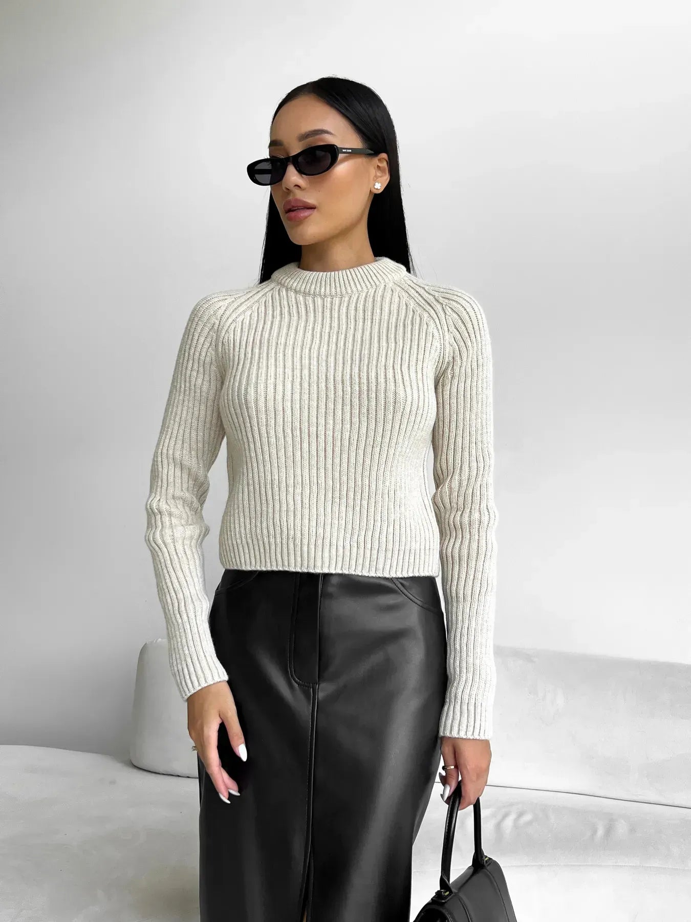 Kvinde i hvid striktrøje og sort lædernederdel, iført solbriller, står foran en lys baggrund. Fashionabelt outfit, moderne stil, minimalistisk look.