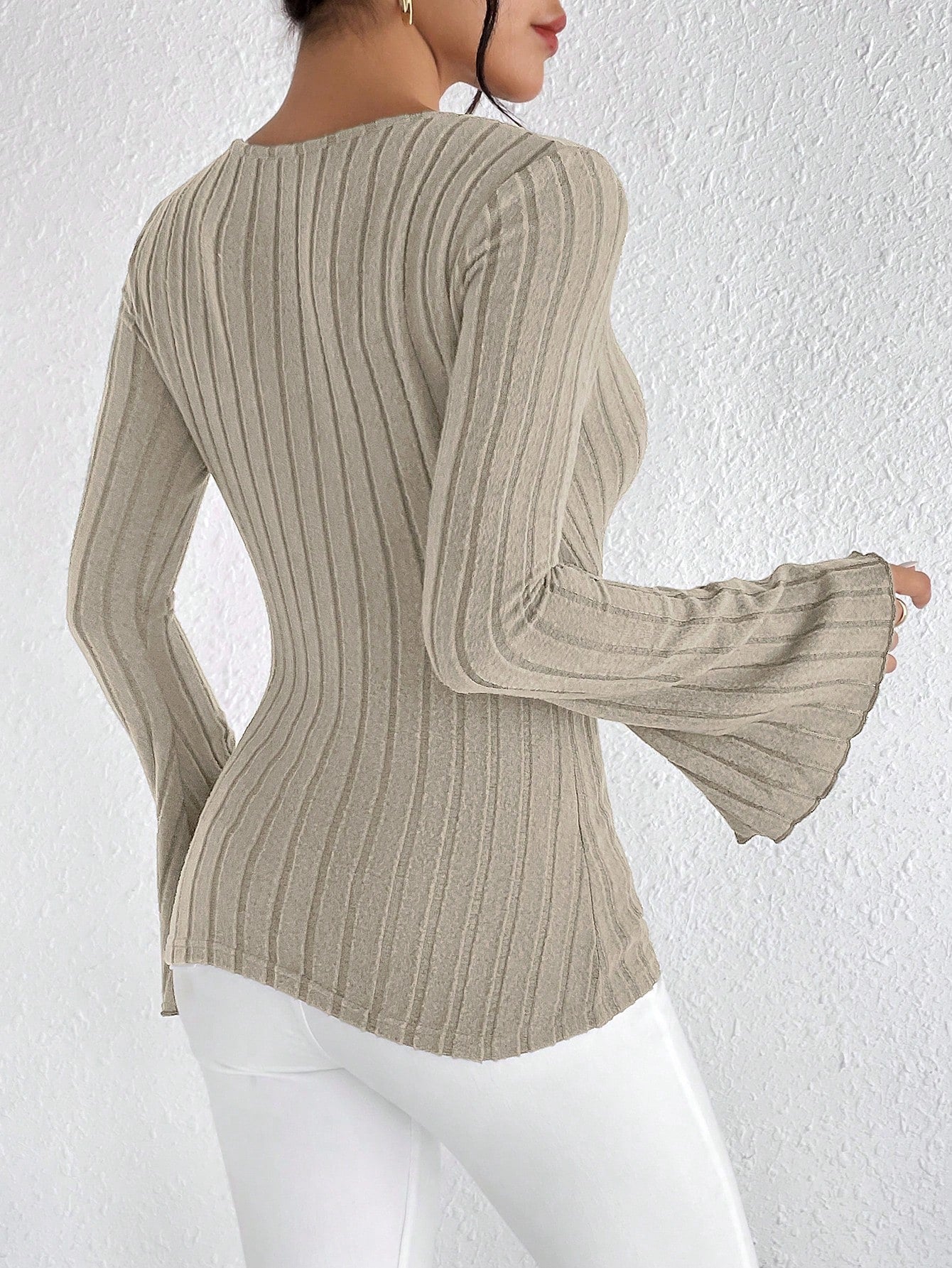 Kvinde iført beige ribstrikket trøje med klokkeærmer og hvide bukser, set bagfra mod hvid væg. Elegant og moderne mode.